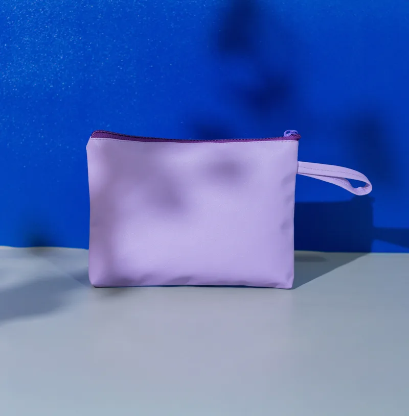 Pouch pipih warna ungu muda dengan ritsleting di atas dan tali pegangan ungu di sisi kanan, di atas latar biru dan abu-abu