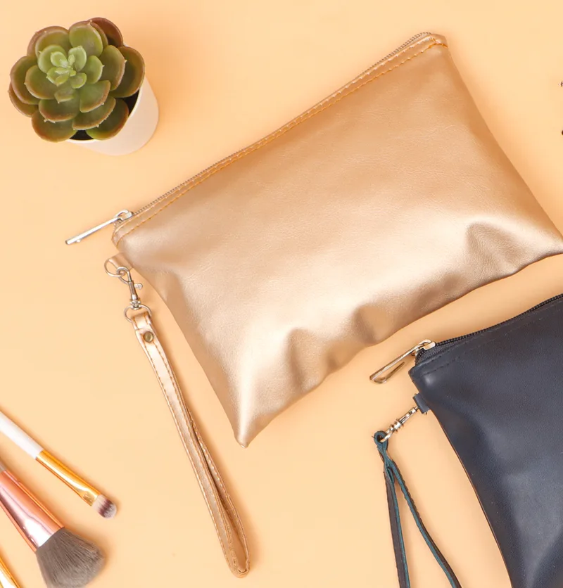Pouch berwarna emas dengan ritsleting perak dan tali tangan berpengait, di samping pouch biru tua, tanaman sukulen dalam pot, kacamata, dan kuas rias di atas latar oranye muda