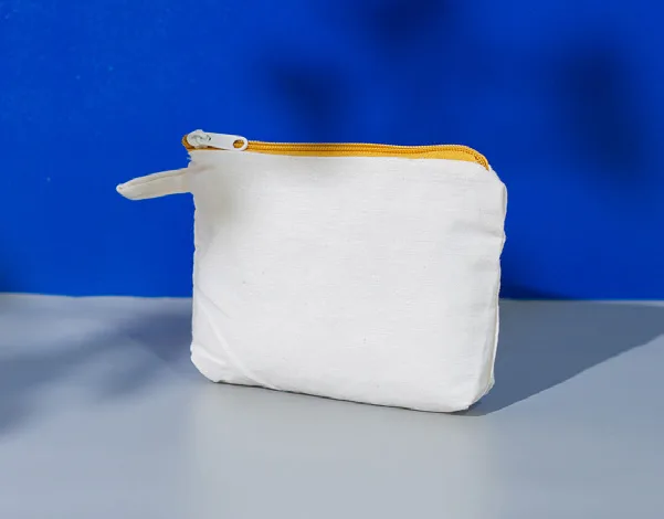 Pouch kapal putih polos dengan ritsleting kuning dan tali kecil di sisi kiri, berdiri di atas meja abu-abu dengan latar dinding biru