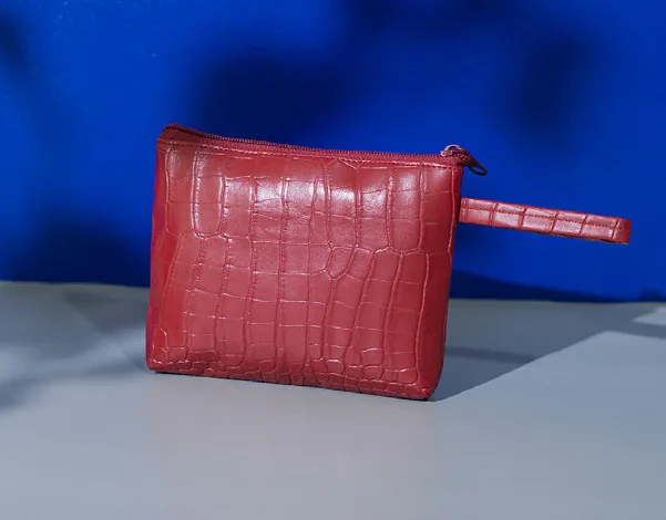 Pouch kapal merah bertekstur motif croco dengan ritsleting merah dan tali tangan bermotif sama, berdiri di atas meja abu-abu dengan latar dinding biru
