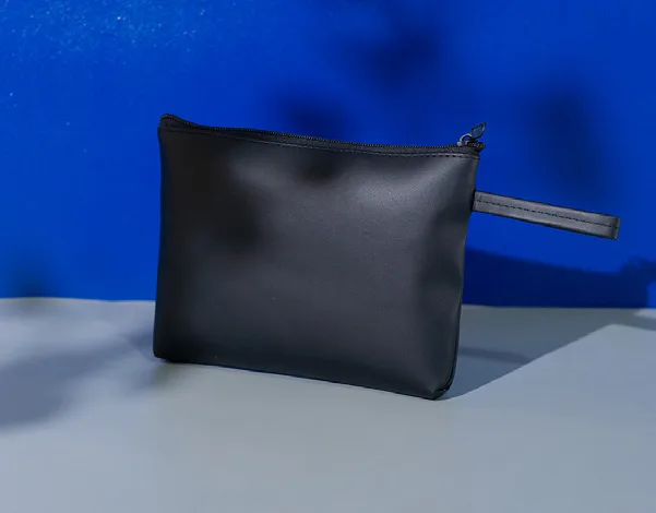 Pouch kapal hitam polos bertekstur doff dengan ritsleting hitam dan tali tangan di sisi kanan, di atas meja abu-abu dengan latar dinding biru