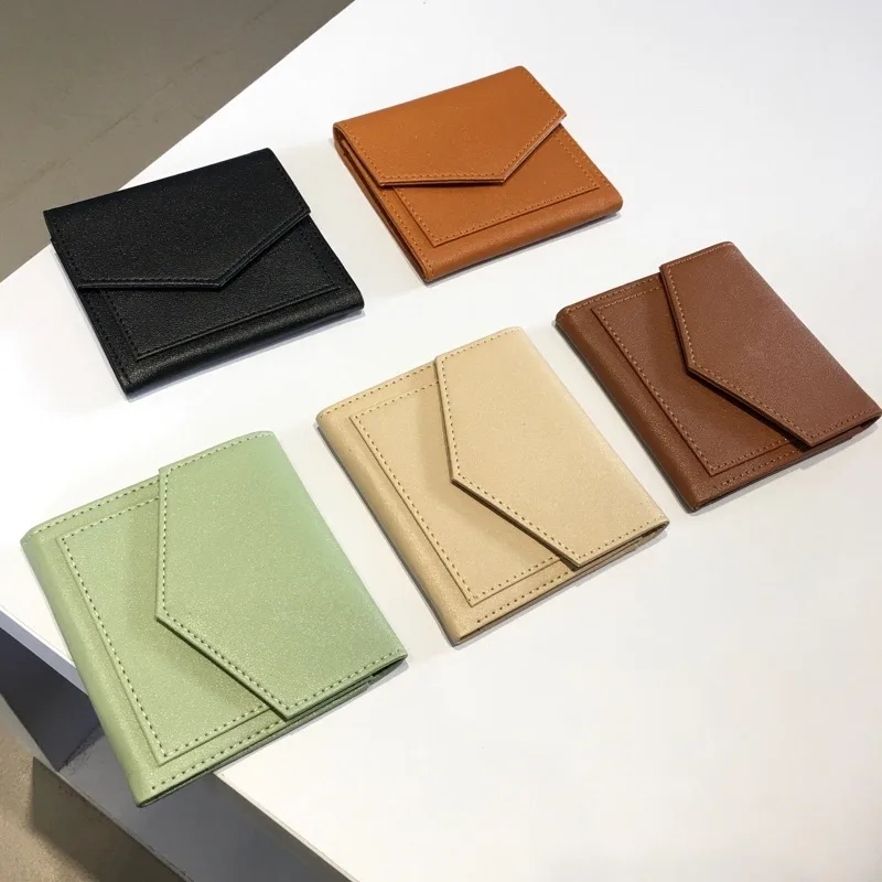 Lima dompet lipat bertutup amplop berwarna hitam, oranye, cokelat tua, hijau mint, dan krem tertata di atas meja putih