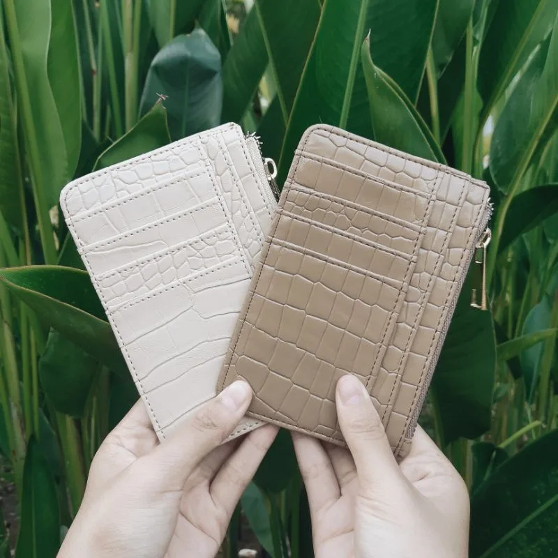 Dua tangan memegang dua dompet kartu bertekstur croco dengan ritsleting di sisi kanan, satu berwarna krem dan satu berwarna cokelat keabu-abuan, dengan latar daun hijau