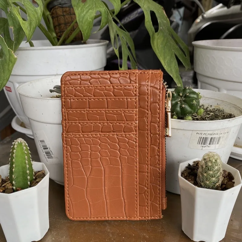 Dompet kartu bertekstur croco berwarna oranye kecokelatan dengan beberapa slot kartu dan ritsleting emas di sisi kanan, berdiri di antara pot tanaman kaktus berwarna putih