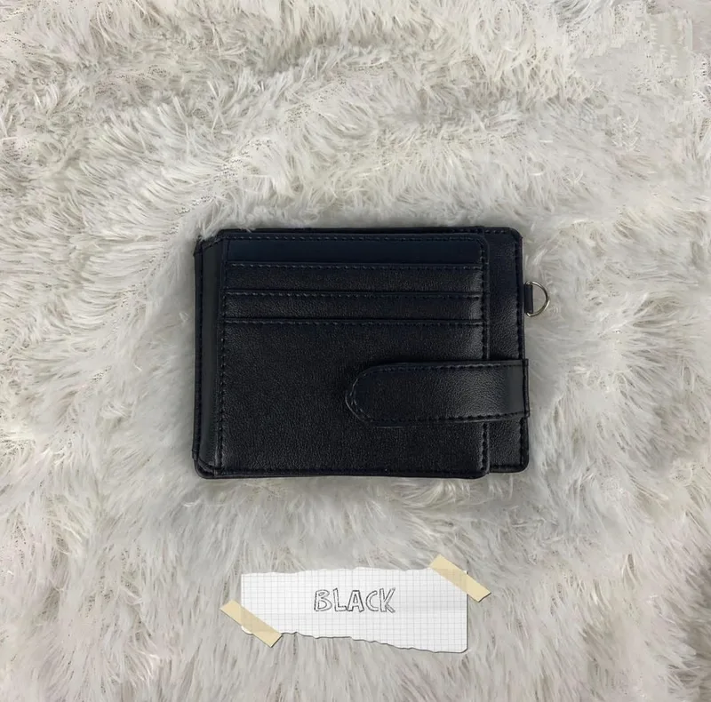 Card holder hitam dengan beberapa slot kartu, tali pengunci, dan ring logam kecil di sisi kanan, diletakkan di atas kain berbulu putih dengan label kertas bertulisan BLACK