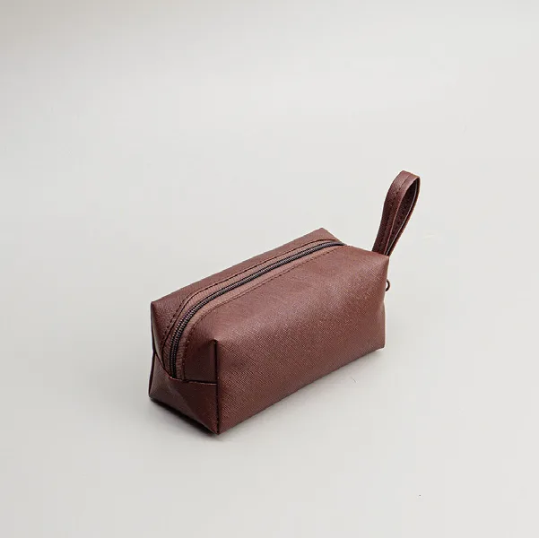 Pouch kubus kecil berwarna cokelat tua dengan ritsleting dan tali di ujung, difoto miring di atas latar abu-abu muda