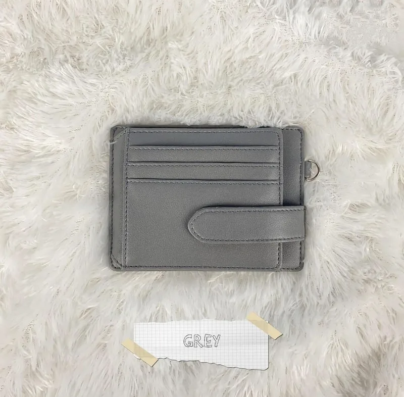 Card holder abu-abu dengan beberapa slot kartu dan tali pengunci berkancing, diletakkan di atas bulu putih dengan label kertas bertuliskan GREY