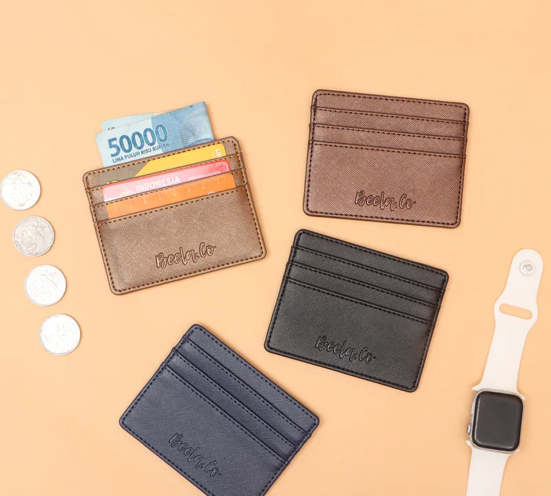 Empat card holder berwarna cokelat keemasan, cokelat tua, hitam, dan biru dongker dengan tulisan Beela.Co yang diemboss, satu di antaranya berisi kartu dan uang lembaran, di atas latar oranye muda bersama koin dan jam tangan