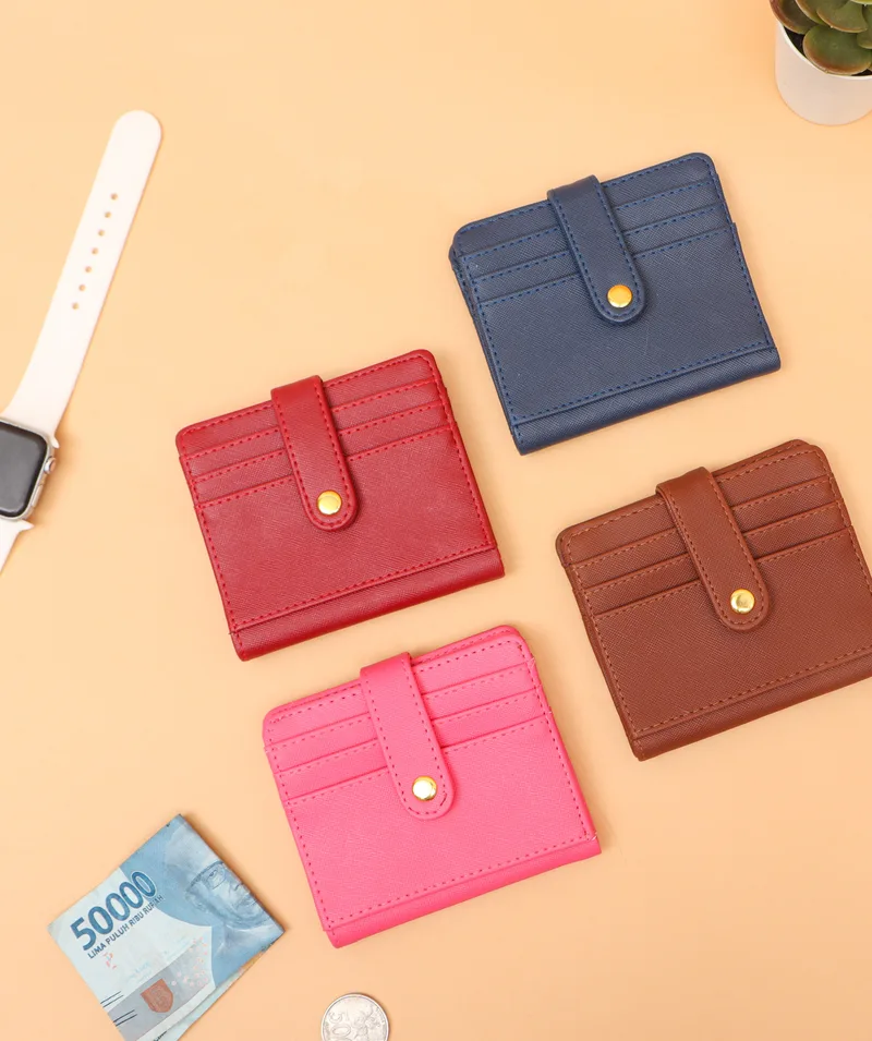 Empat card holder lipat dengan tali berkancing emas berwarna biru dongker, merah, cokelat, dan merah muda di atas latar oranye muda bersama jam tangan dan uang lembaran