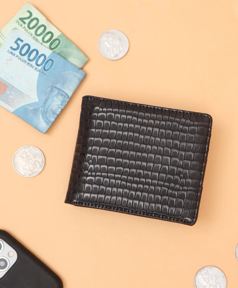 Dompet lipat hitam bertekstur motif kulit buaya di atas latar oranye muda, dikelilingi uang lembaran, koin, dan sebagian ponsel