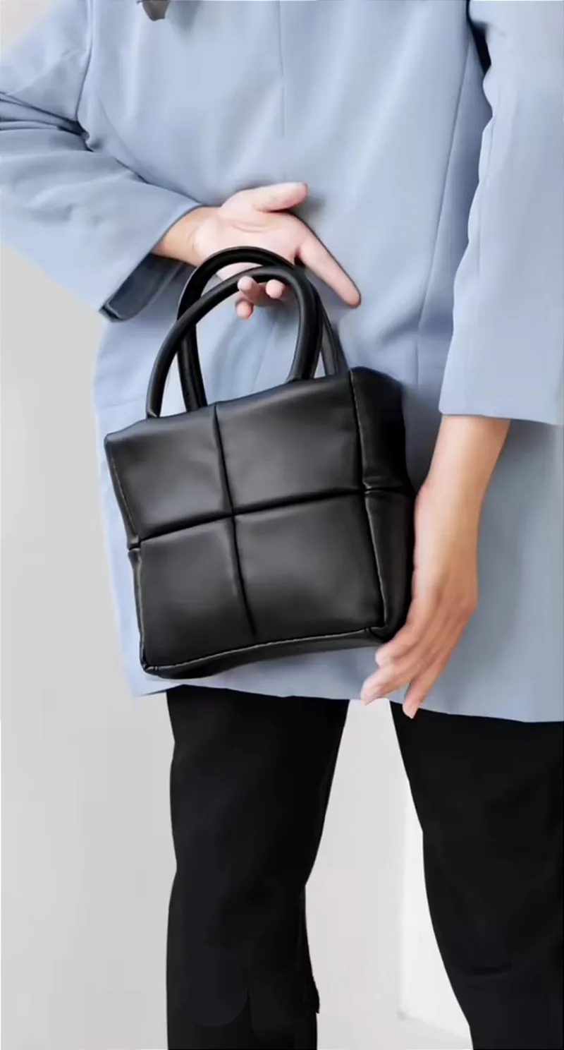 Tas tangan hitam kotak dengan pola jahitan empat panel dan dua pegangan bulat, dipegang di depan blazer biru muda dan celana hitam