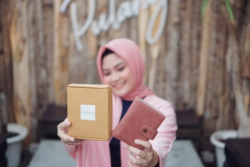 Perempuan berhijab merah muda memegang kotak karton cokelat berlabel Beela.co di tangan kanan dan dompet merah muda di tangan kiri