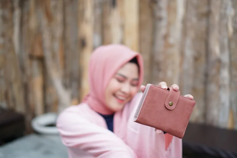 Perempuan berhijab merah muda tersenyum sambil menjulurkan dompet lipat berwarna merah muda dengan tali kancing jepret ke depan kamera