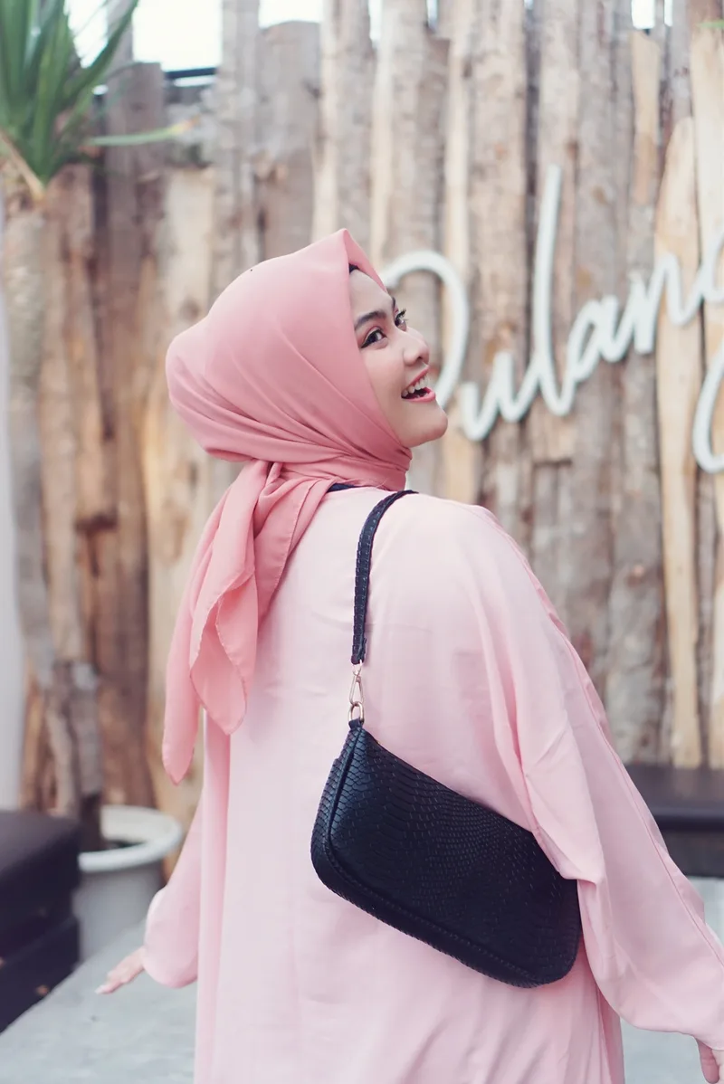 Perempuan berhijab dan gamis merah muda tertawa menoleh ke belakang dengan tas bahu hitam bertekstur sisik tersampir di bahunya, berlatar pagar kayu