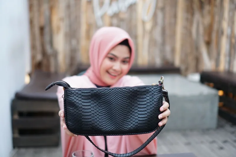 Perempuan berhijab merah muda tersenyum sambil menyodorkan tas bahu hitam bertekstur sisik dengan ritsleting dan pengait emas ke arah kamera
