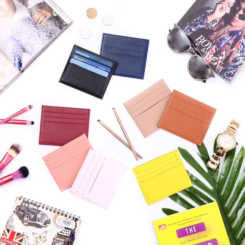 Foto dari atas: card holder dalam warna hitam, biru dongker, merah marun, peach, merah muda, krem, cokelat tan, dan kuning, ditata bersama kuas rias, jam tangan emas, kacamata, dan daun palem di latar putih