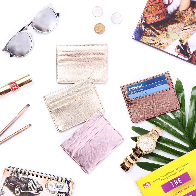 Foto dari atas: card holder metalik berwarna emas muda, champagne, merah muda, dan perunggu, salah satunya berisi kartu, ditata bersama kacamata, lipstik, jam tangan emas, dan daun palem di latar putih