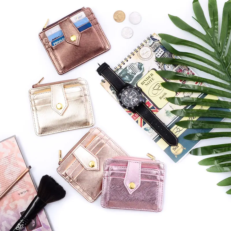 Foto dari atas: empat dompet kartu beritsleting berwarna perunggu, emas, champagne, dan merah muda metalik dengan tab berkancing, ditata bersama jam tangan, buku catatan, kuas rias, dan daun palem di latar putih