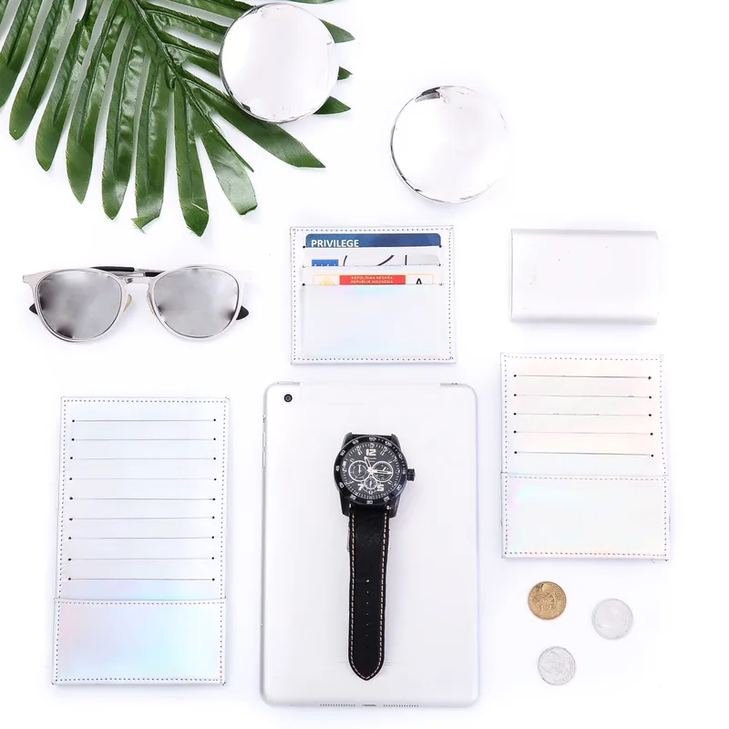 Foto dari atas: tiga card holder perak berkilau pelangi, salah satunya berisi kartu, ditata bersama tablet, jam tangan hitam, kacamata hitam, koin, dan daun palem di atas latar putih