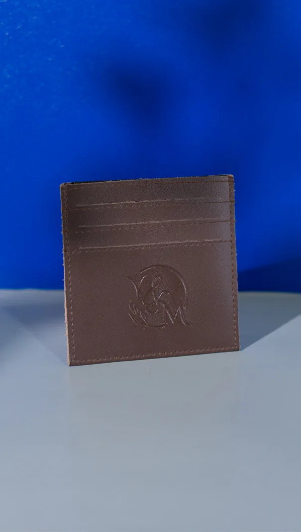 Card holder kulit cokelat dengan tiga slot kartu dan emboss monogram inisial di bagian bawah, berdiri di atas meja dengan latar biru