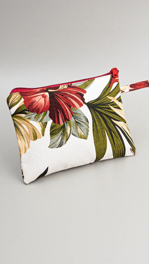 Pouch kanvas bermotif bunga merah besar dan daun hijau di atas dasar putih dengan ritsleting merah di bagian atas, diletakkan miring di atas alas abu-abu