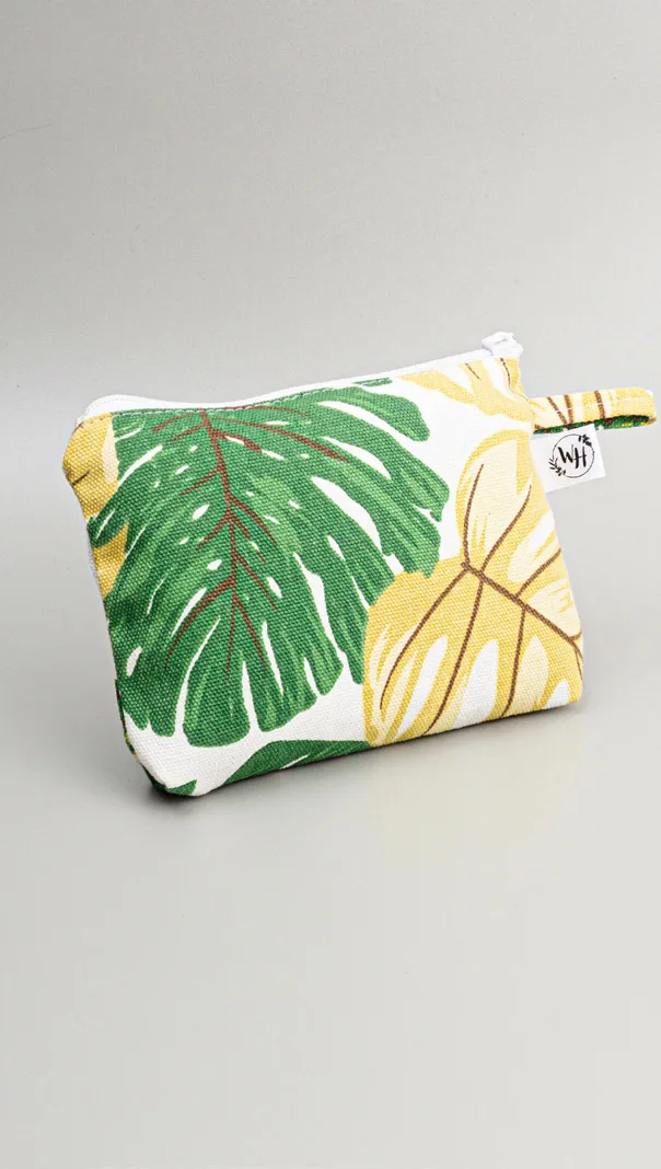 Pouch kanvas bermotif daun monstera hijau dan daun kuning di atas dasar putih, dengan ritsleting di bagian atas dan tali kecil di sisi kanan, diletakkan di atas alas abu-abu
