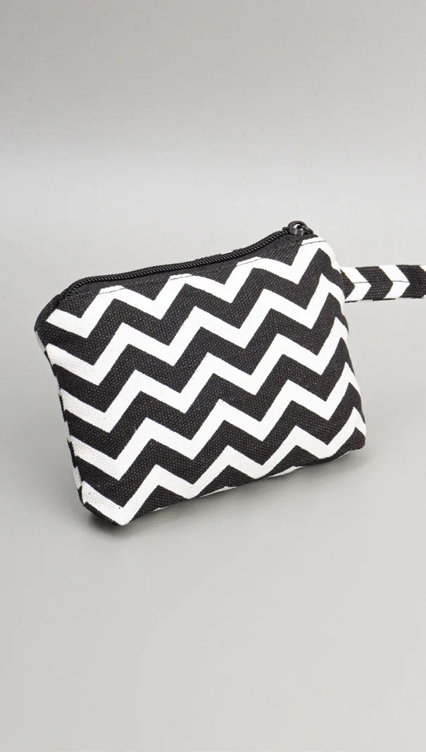 Pouch kanvas bermotif zigzag hitam putih dengan ritsleting di bagian atas dan tali kecil bermotif sama di sisi kanan, di atas alas abu-abu