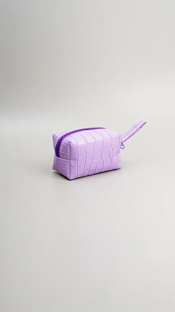 Pouch kecil berbentuk kubus berwarna ungu muda dengan tekstur kotak menyerupai kulit buaya, ritsleting senada, dan tali pegangan di sisi kanan, di atas alas abu-abu