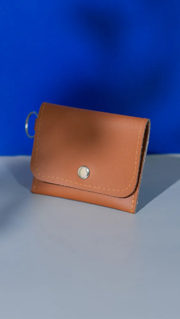 Card holder cokelat dengan penutup lipat berkancing logam dan cincin gantungan kunci di sisi atas, difoto di atas latar biru