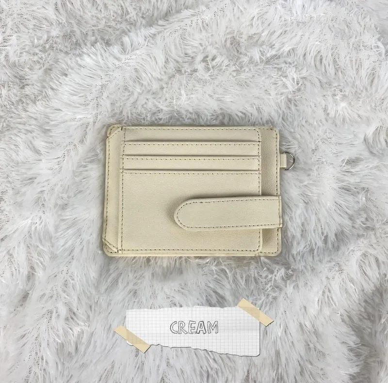 Card holder Side Flip warna cream dengan tiga slot kartu dan tali pengunci, diletakkan di atas karpet bulu putih dengan label kertas bertuliskan CREAM