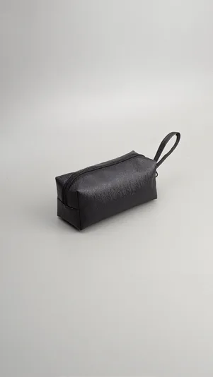 Pouch kubus hitam kecil bertekstur halus dengan ritsleting dan tali tangan, diletakkan di atas latar abu-abu muda