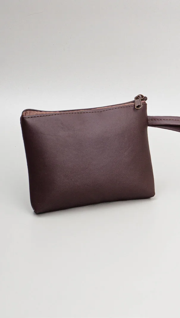 Pouch pipih model kapal berbahan kulit warna cokelat tua dengan ritsleting dan tali pegangan, difoto di atas meja abu-abu