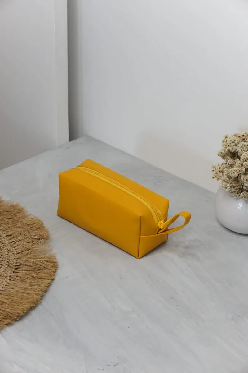 Pouch kubus kulit sintetis warna kuning dengan ritsleting senada di atas meja beton abu-abu, di sampingnya alas anyaman dan vas bunga kering