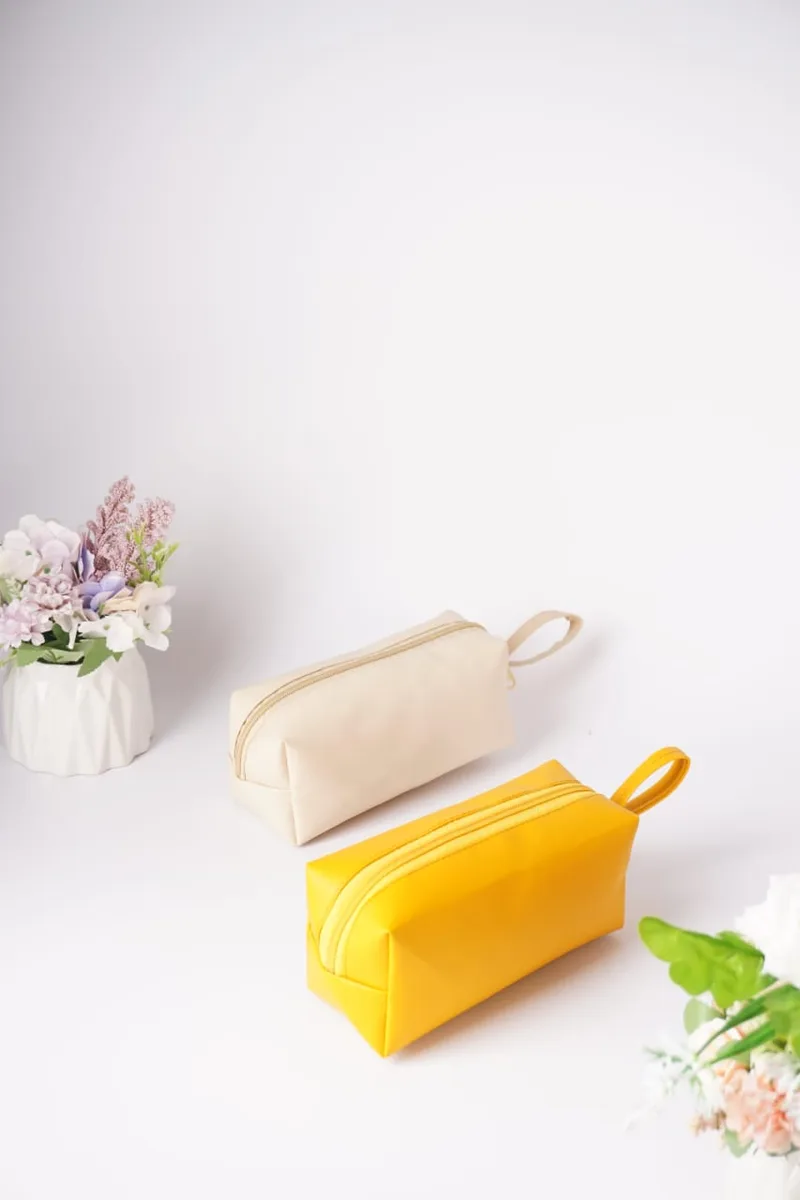 Dua pouch kubus kulit sintetis, satu warna krem dan satu warna kuning, dengan ritsleting senada dan tali pegangan kecil, di atas meja putih dengan bunga