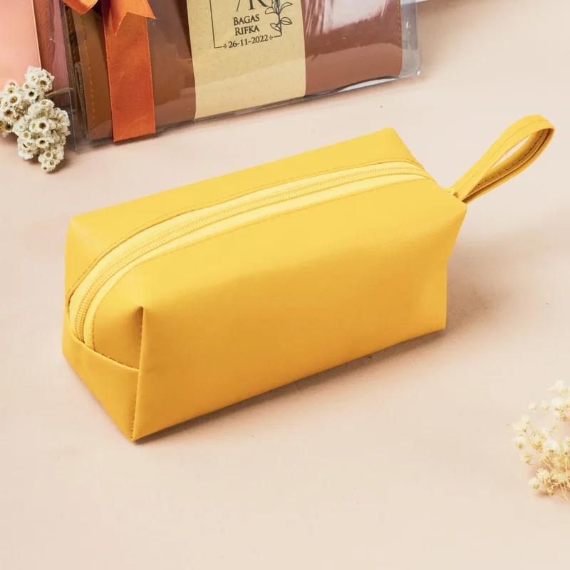 Pouch kubus kulit sintetis warna kuning di depan, di belakangnya set souvenir pernikahan yang sudah dikemas dalam plastik bening dengan pita oranye dan label bertuliskan nama pasangan Bagas dan Rifka serta tanggal 26-11-2022