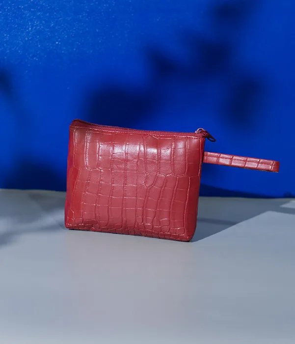 Pouch merah bertekstur motif kulit buaya dengan ritsleting di atas dan tali pegangan bermotif sama di sisi kanan, berdiri di atas alas abu-abu dengan latar biru dan bayangan daun