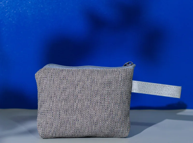 Pouch kain bertekstur anyaman abu-abu dengan ritsleting abu-abu di atas dan tali pegangan abu-abu muda di sisi kanan, berdiri di atas alas abu-abu dengan latar biru