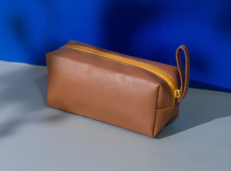 Pouch kubus warna cokelat dengan ritsleting kuning keemasan dan tali pegangan di sisi kanan, di atas latar biru