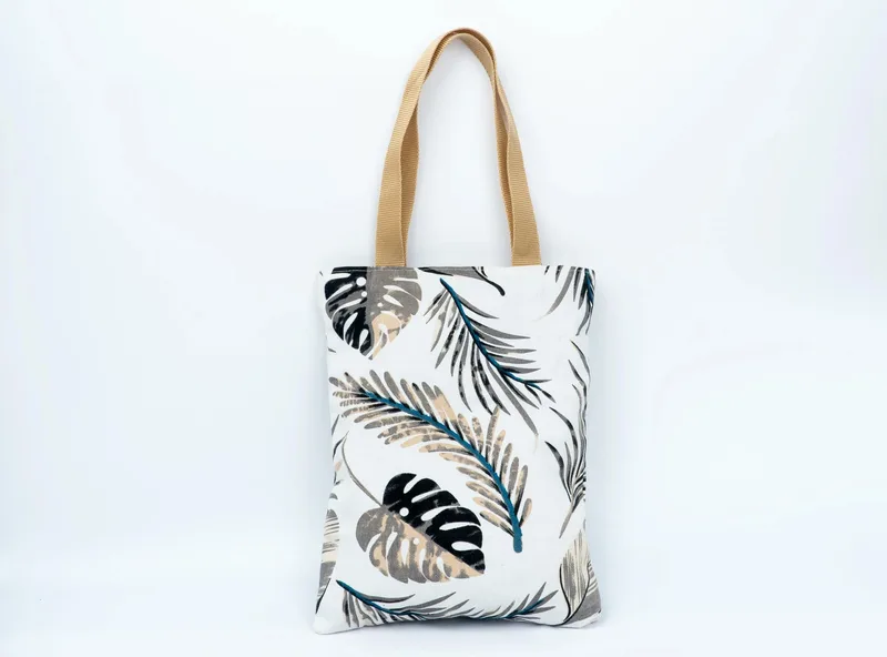 Tote bag kanvas putih bermotif daun tropis hitam dan krem dengan tali pegangan warna cokelat muda