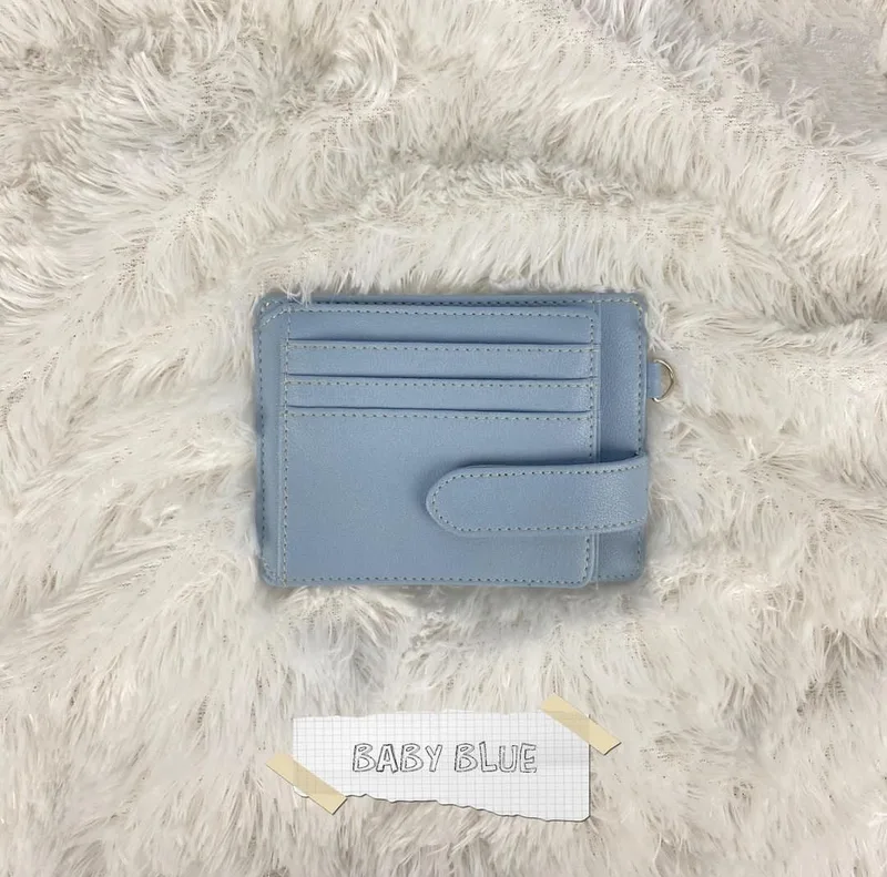 Card holder berwarna biru muda dengan slot kartu bertingkat dan tali pengunci berkancing di atas karpet bulu putih, dengan label kertas bertuliskan Baby Blue