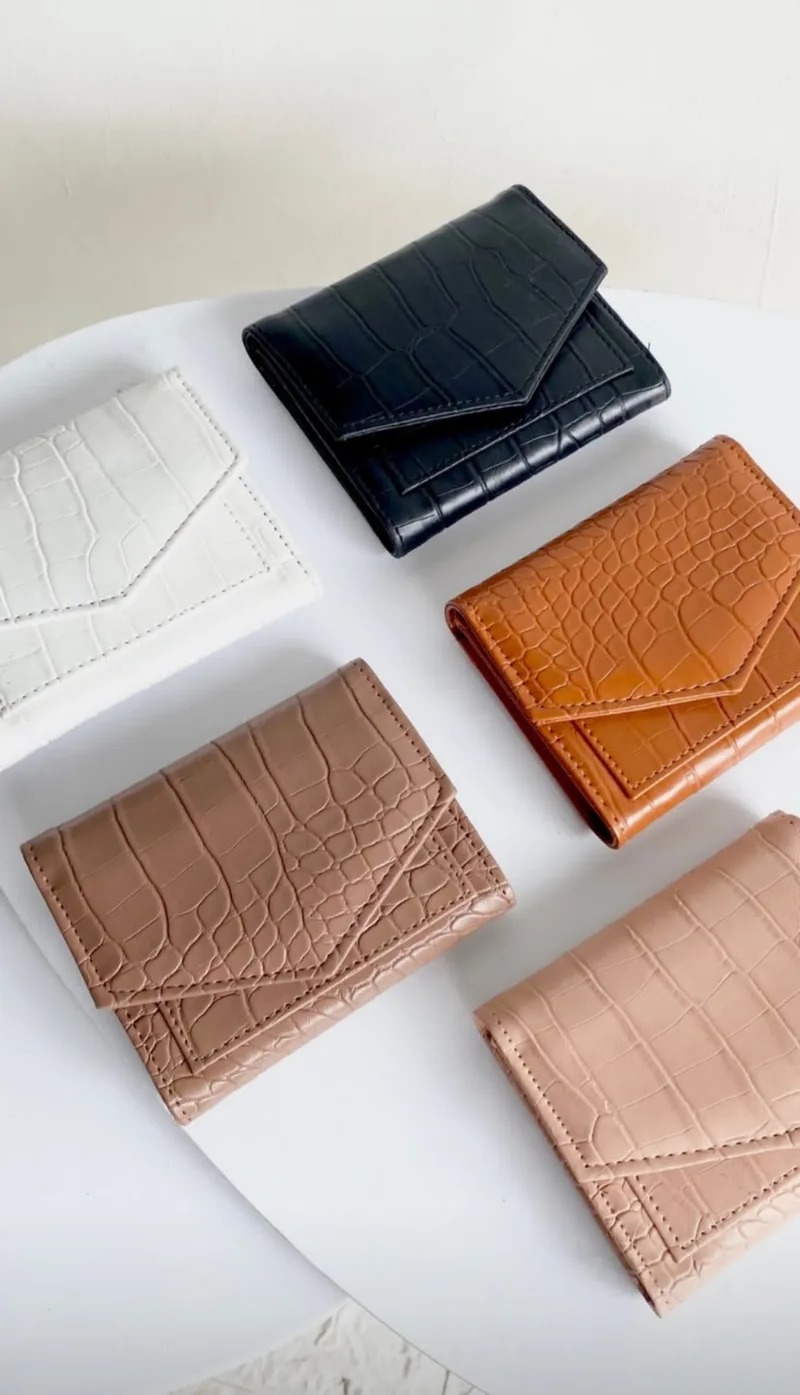 Enam dompet lipat kecil bertekstur motif croco dalam warna putih, hitam, oranye kecokelatan, cokelat susu, dan merah muda, ditata berjajar di atas kain putih