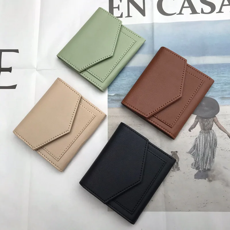 Empat dompet lipat kecil polos berpenutup amplop dalam warna hijau sage, krem, cokelat kemerahan, dan hitam, diletakkan di atas lembar kertas bergambar pantai