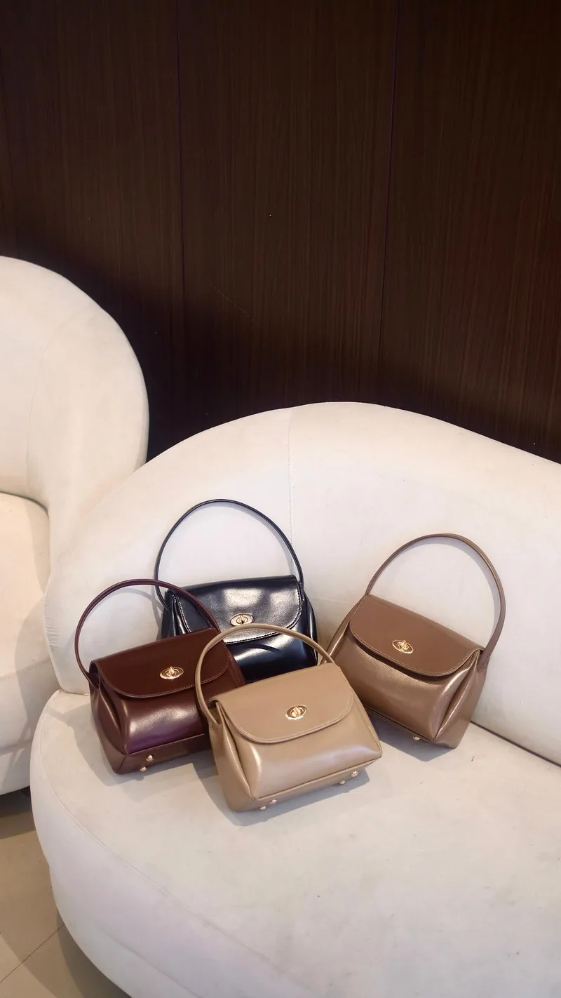 Empat sling bag mungil berbahan mengilap warna hitam, cokelat tua, krem, dan cokelat muda dengan pegangan atas dan kancing putar logam, ditata di kursi putih