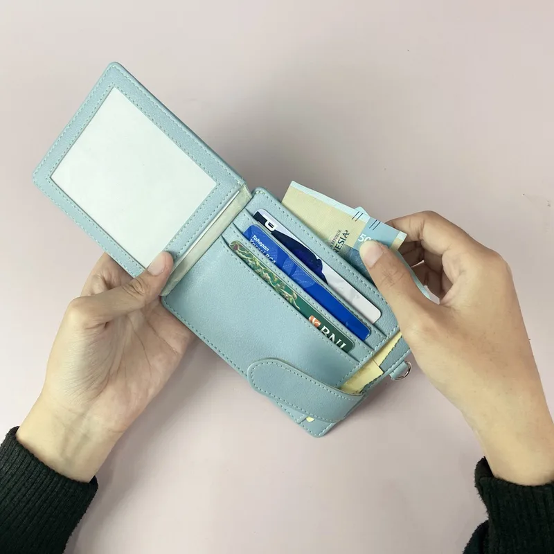 Dua tangan membuka card holder biru muda yang berisi beberapa kartu dan uang kertas, dengan jendela transparan untuk kartu identitas di sisi kiri