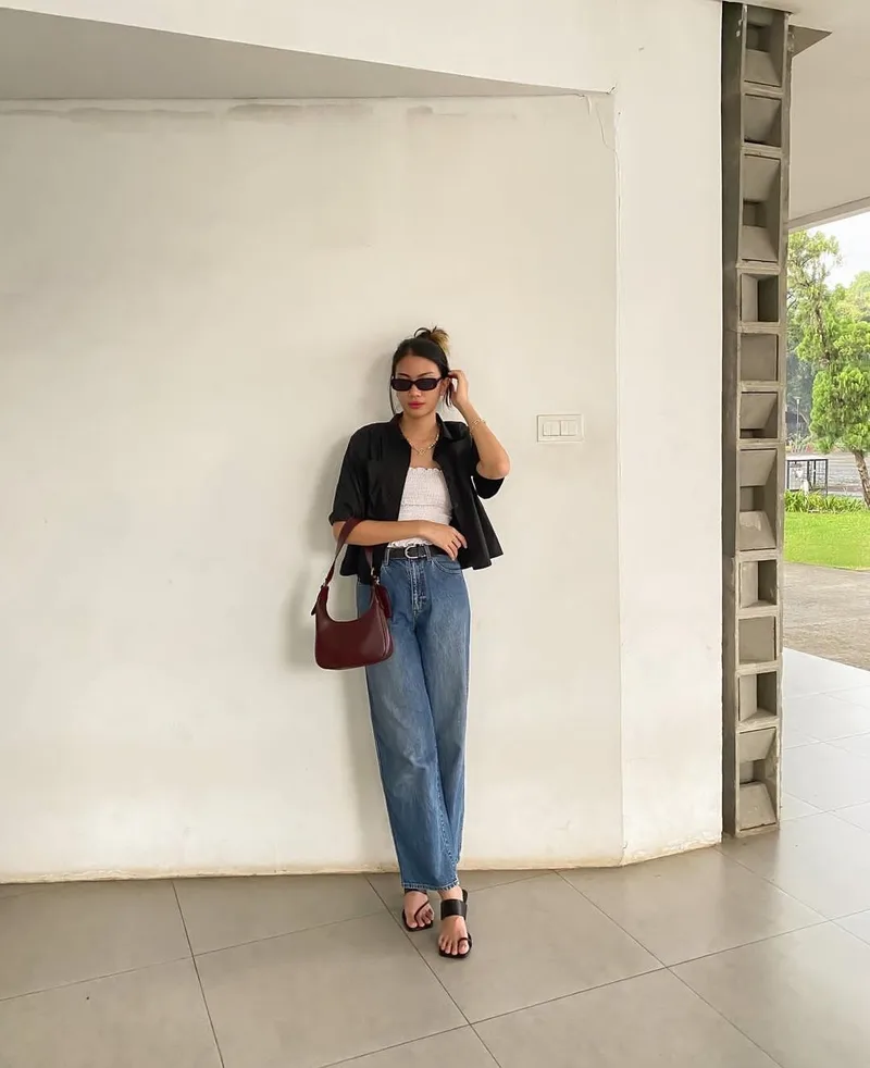 Wanita berkacamata hitam, kemeja hitam, atasan putih, dan celana jeans biru longgar bersandar di dinding putih sambil memegang tas bahu merah marun di samping pinggang
