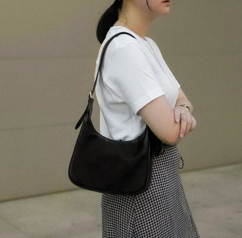 Wanita berkaus putih dan rok kotak-kotak hitam putih berdiri dengan tangan terlipat, memakai tas bahu hitam berbentuk melengkung yang talinya digantung di bahu kanan sehingga tas berada di bawah ketiak