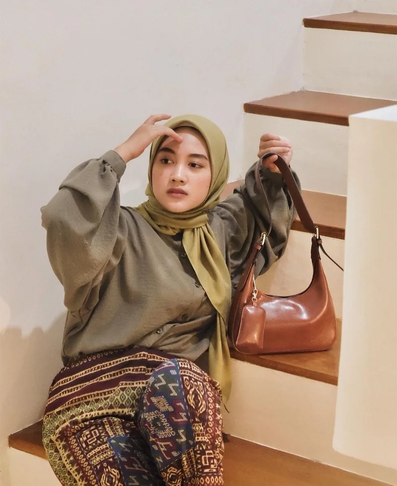 Wanita berhijab hijau zaitun dan kemeja hijau keabuan duduk di anak tangga kayu, memegang tali tas bahu cokelat tan dengan pouch kecil, mengenakan celana bermotif tenun