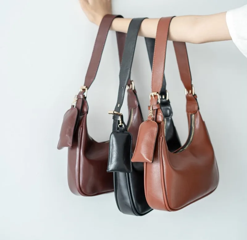 Tiga tas bahu melengkung berwarna merah marun, hitam, dan cokelat digantung bersama di lengan seorang wanita, masing-masing dengan pouch kecil di pengait tali
