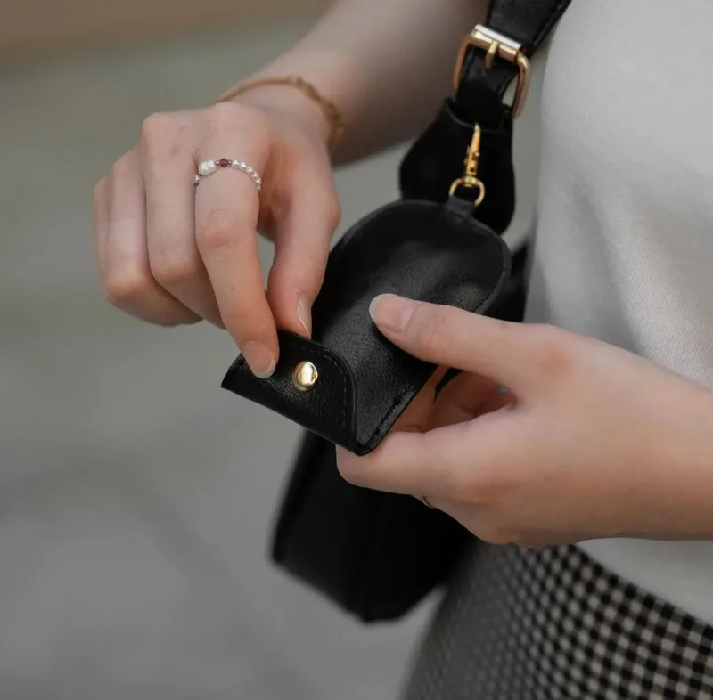 Tangan wanita bercincin membuka kancing logam emas pada pouch kecil hitam yang tergantung di pengait tali tas bahu hitam