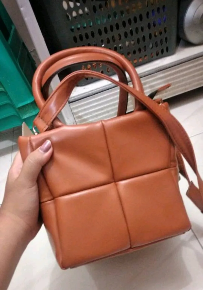 Tangan berkuteks memegang tas tangan cokelat karamel berpanel kotak dengan dua pegangan bulat pendek dan tali panjang yang dilipat di atasnya, di dalam ruangan
